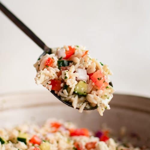 Large serving spoon filled with Mediterranean orzo salad hovering over a large bowl filled with the salad.