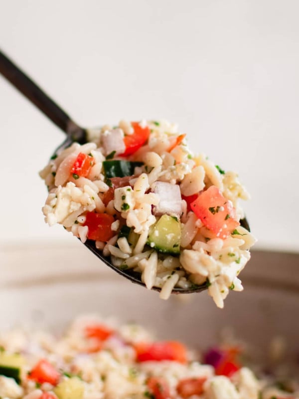 Large serving spoon filled with Mediterranean orzo salad hovering over a large bowl filled with the salad.