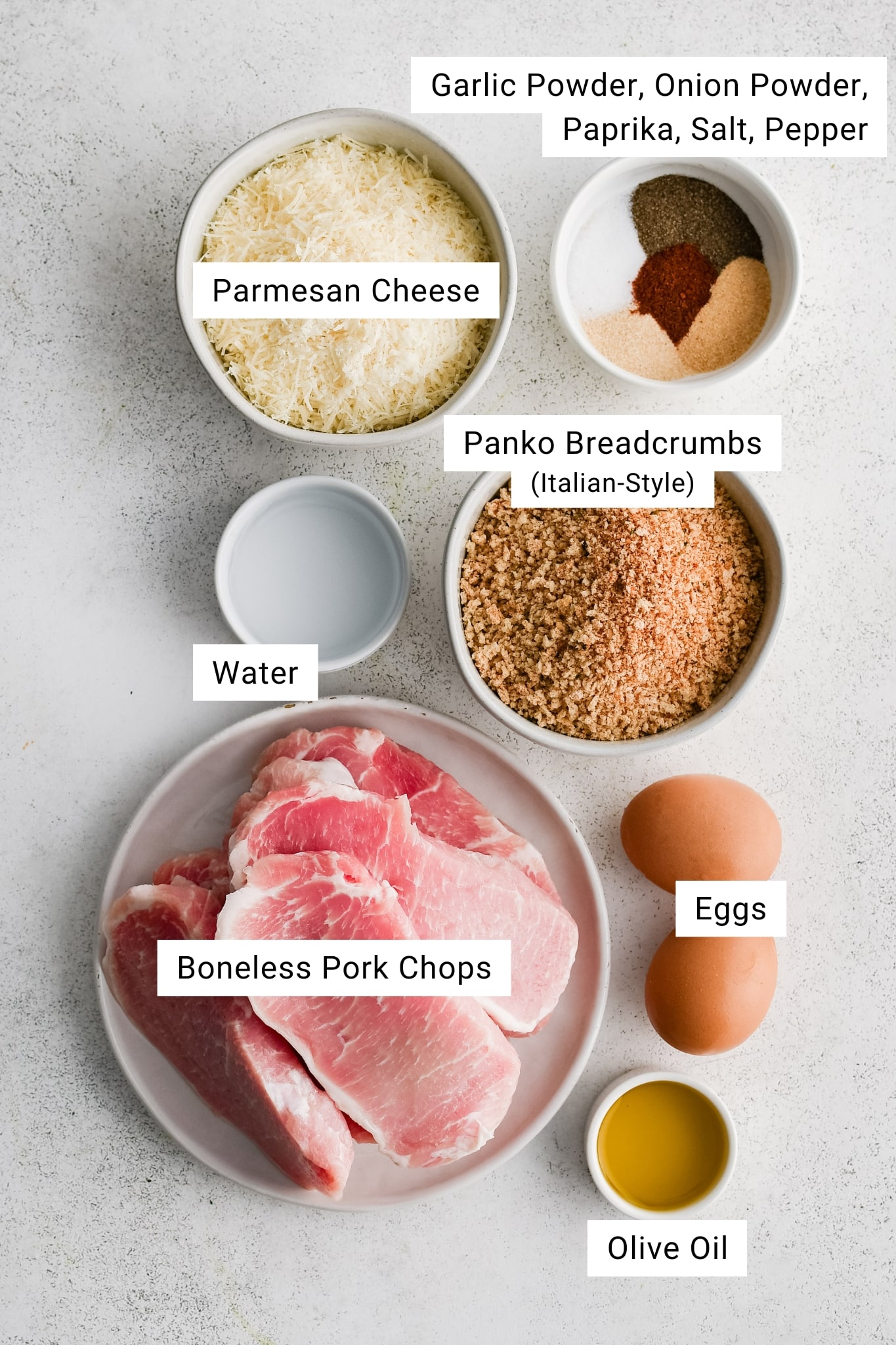 Overhead view of boneless pork chops, shredded Parmesan, Italian style panko breadcrumbs, eggs, olive oil, water, and small bowls of garlic powder, onion powder, paprika, salt, and pepper arranged on a light surface.