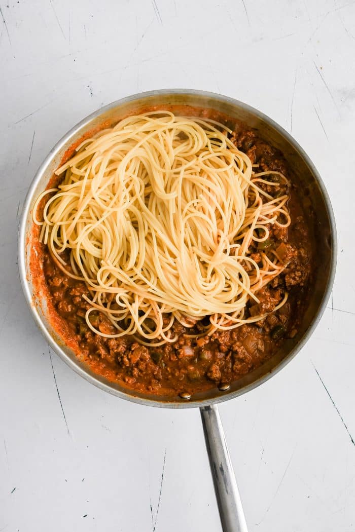 Al Dente spaghetti noodles added to a large stainless steel skillet filled with meaty tomato sauce.