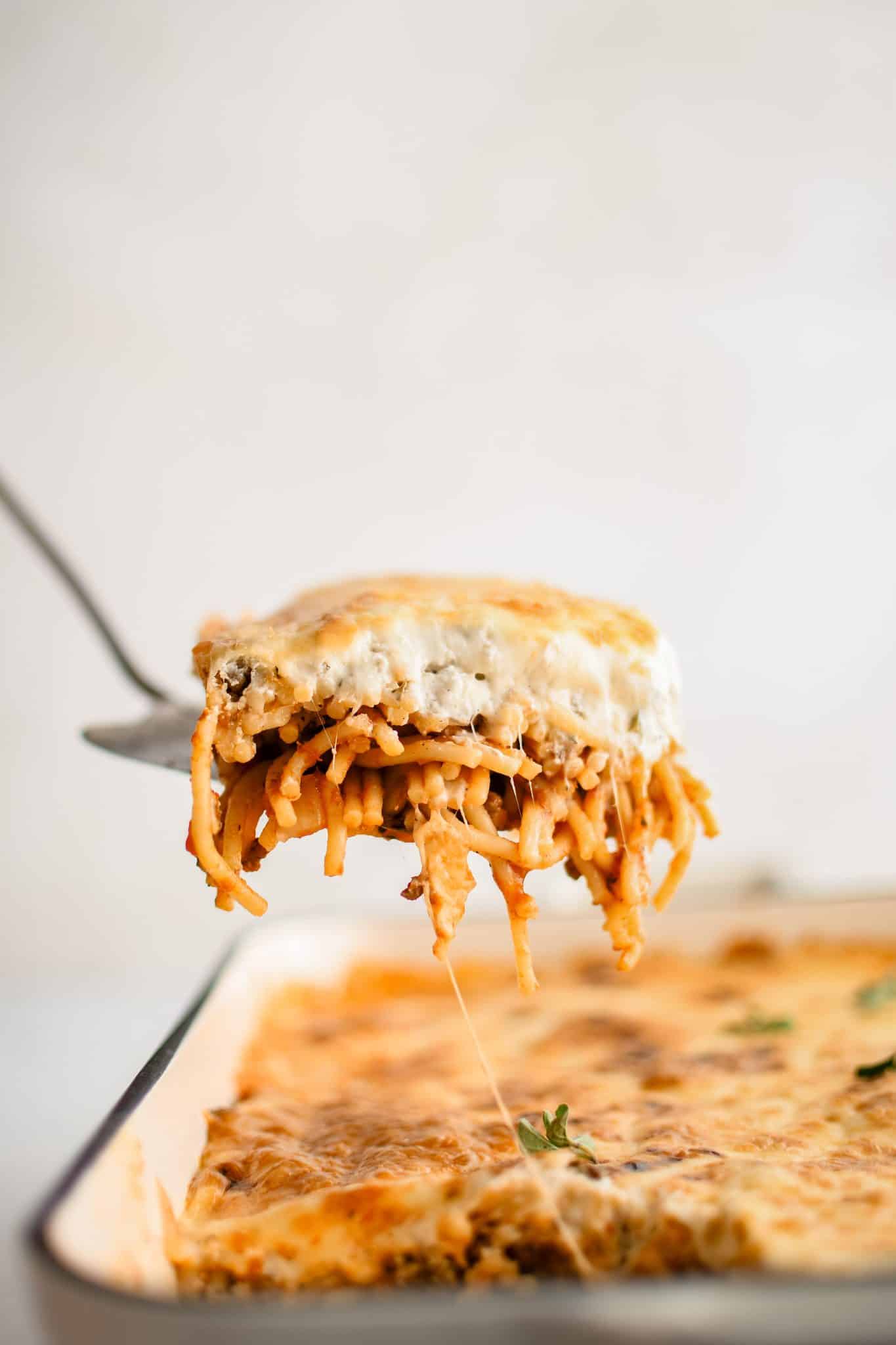 Metal Spatula with a slice of spaghetti casserole showing the layer of spaghetti sauce covered noodles and cream cheese layer floating above a large casserole dish filled with spaghetti casserole