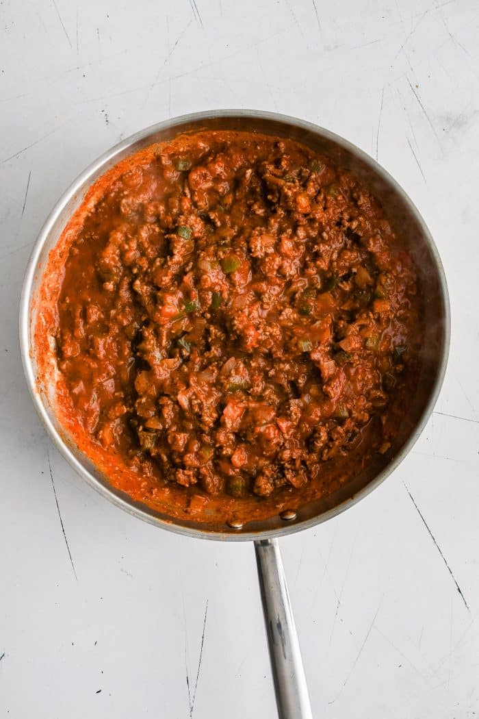 Large stainless steel skillet filled with ground beef, diced onion and green bell peppers mixed in tomato sauce.