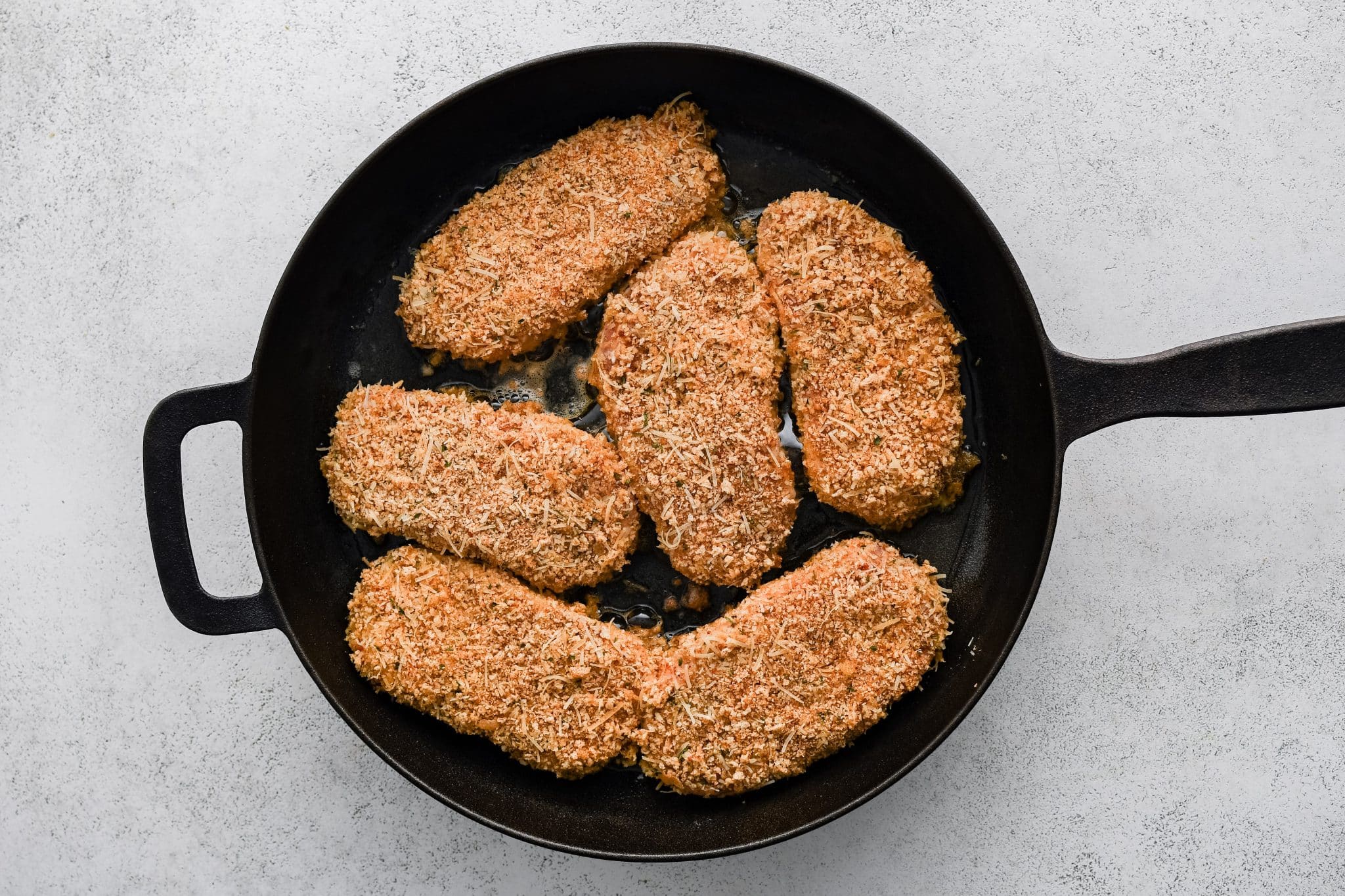 Six breaded pork chops arranged in a large cast iron skillet with a light sizzle and pale golden spots.