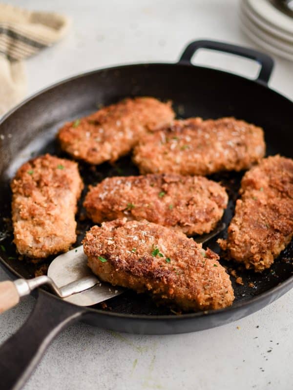 Cast iron skillet with six golden Parmesan crusted pork chops, one lifted slightly with a metal spatula, crisp panko crust and parsley flecks visible.