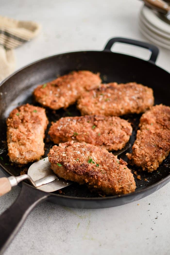 Cast iron skillet with six golden Parmesan crusted pork chops, one lifted slightly with a metal spatula, crisp panko crust and parsley flecks visible.