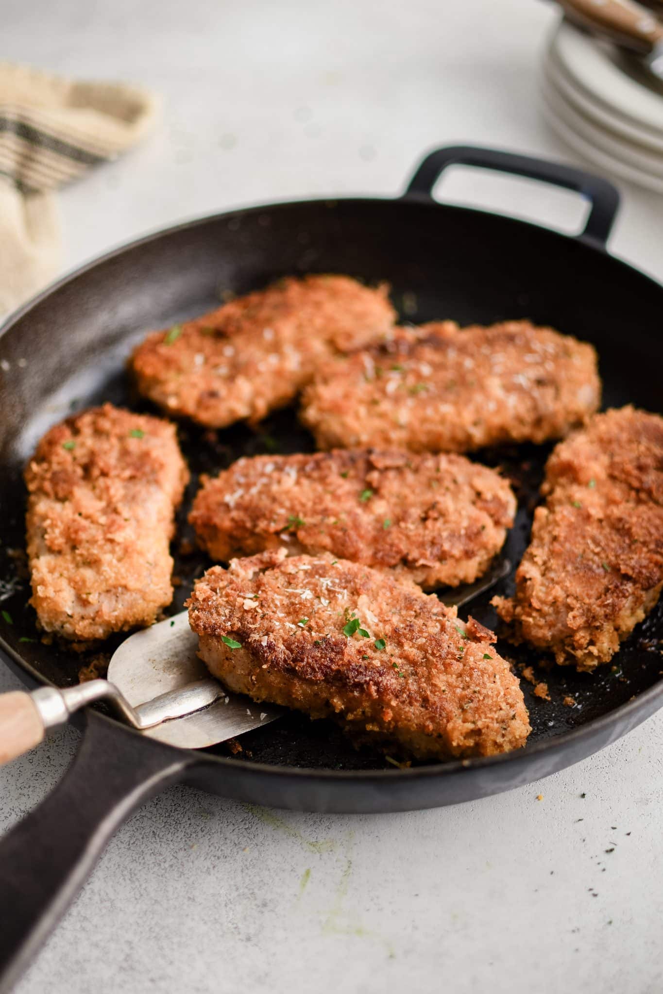Cast iron skillet with six golden Parmesan crusted pork chops, one lifted slightly with a metal spatula, crisp panko crust and parsley flecks visible.