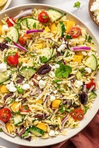 Close-up of Mediterranean orzo salad with tender orzo pasta, crumbled feta cheese, sliced cucumber, cherry tomatoes, yellow bell pepper, red onion, Kalamata olives, fresh parsley, mint, and cracked black pepper in a beige bowl with a rust-colored napkin nearby.