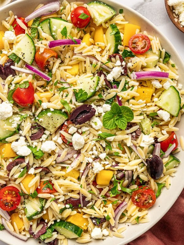 Close-up of Mediterranean orzo salad with tender orzo pasta, crumbled feta cheese, sliced cucumber, cherry tomatoes, yellow bell pepper, red onion, Kalamata olives, fresh parsley, mint, and cracked black pepper in a beige bowl with a rust-colored napkin nearby.