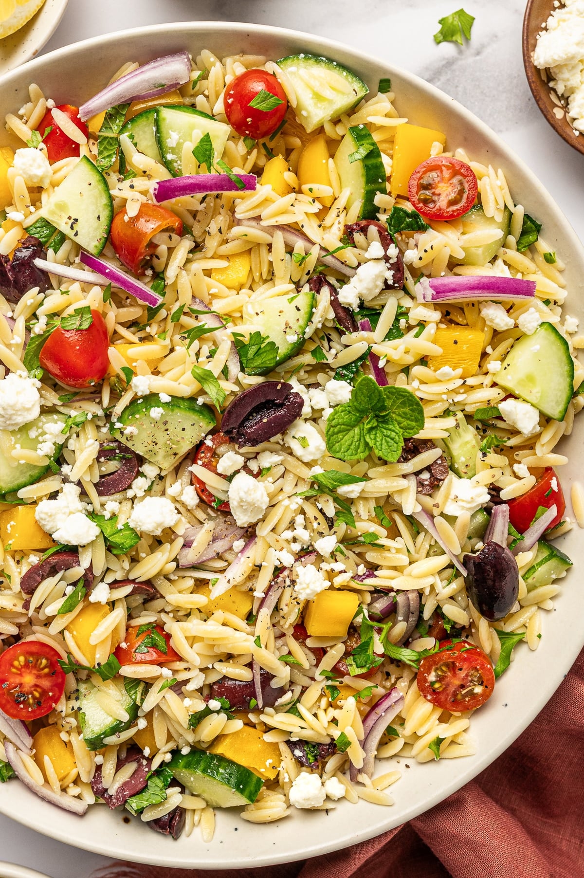 Close-up of Mediterranean orzo salad with tender orzo pasta, crumbled feta cheese, sliced cucumber, cherry tomatoes, yellow bell pepper, red onion, Kalamata olives, fresh parsley, mint, and cracked black pepper in a beige bowl with a rust-colored napkin nearby.