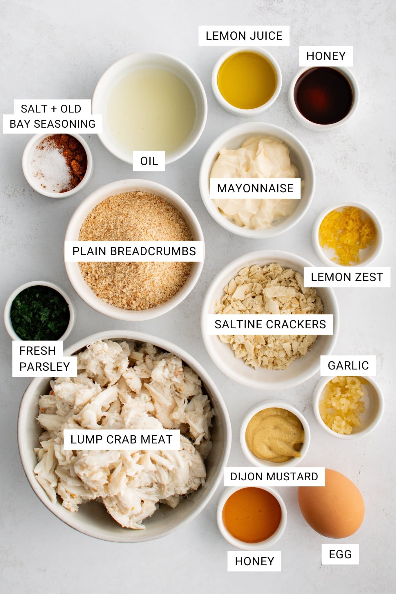 Overhead view of ingredients for homemade crab cakes arranged in white bowls on a light gray surface. The image includes labeled text overlays for lump crab meat in a large white bowl, fresh parsley, plain breadcrumbs, crushed saltine crackers, mayonnaise, Dijon mustard, egg, garlic, lemon zest, lemon juice, oil, honey, and salt with Old Bay seasoning. The ingredients are arranged neatly with white rectangular labels and black text identifying each item.