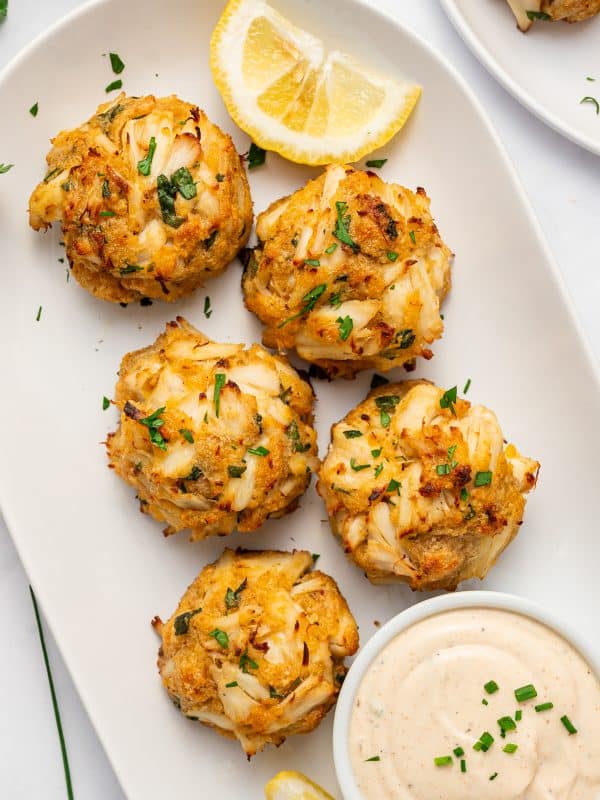 Five golden crab cakes with visible pieces of lump crab meat served on a white oval platter with lemon wedges, chopped parsley, and a small white bowl of creamy sauce garnished with chives, surrounded by fresh parsley, lemon wedges, a fork, and a black and white checkered towel.