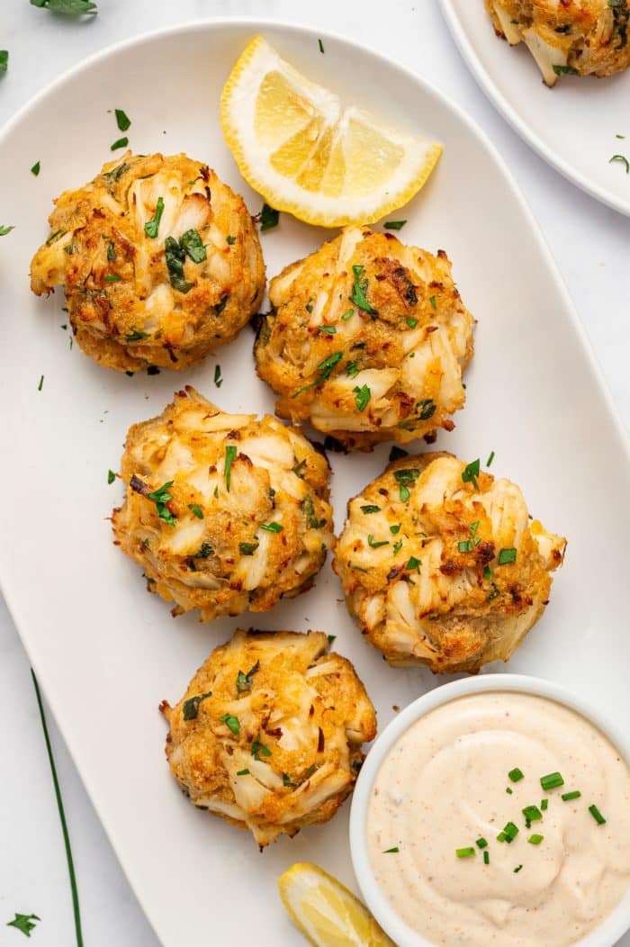 Five golden crab cakes with visible pieces of lump crab meat served on a white oval platter with lemon wedges, chopped parsley, and a small white bowl of creamy sauce garnished with chives, surrounded by fresh parsley, lemon wedges, a fork, and a black and white checkered towel.