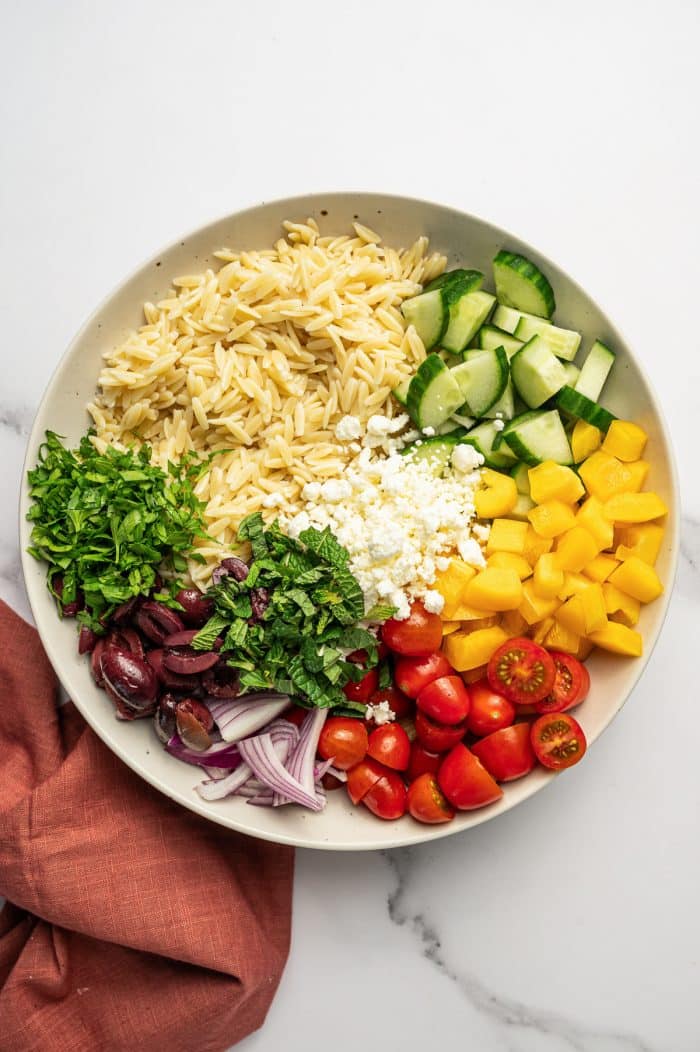 Large beige bowl filled with Mediterranean orzo salad ingredients arranged separately, including cooked orzo, diced cucumber, yellow bell pepper, halved cherry tomatoes, sliced red onion, Kalamata olives, crumbled feta cheese, chopped parsley, and fresh mint on a white marble surface with a rust-colored napkin.