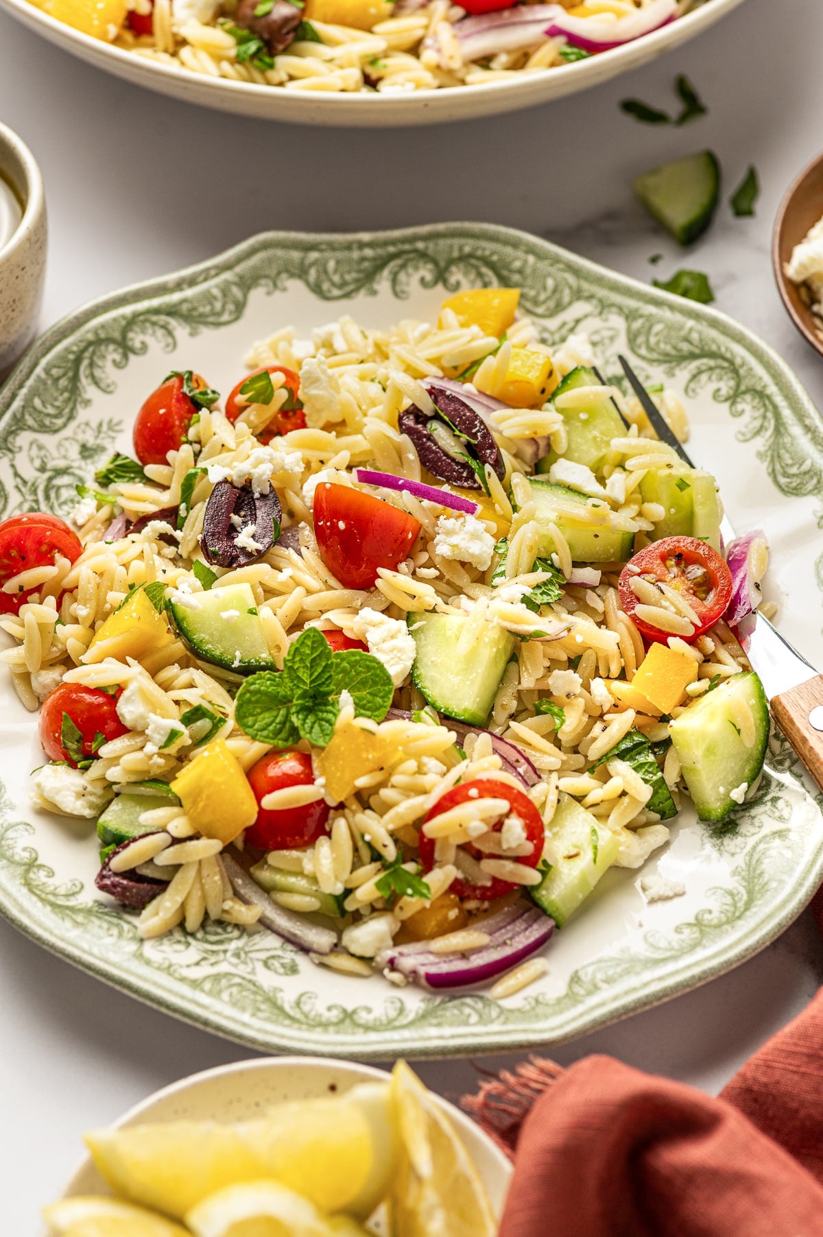Mediterranean orzo salad served on a white plate with a green patterned rim, topped with crumbled feta, fresh mint, cucumber, cherry tomatoes, yellow bell pepper, red onion, Kalamata olives, parsley, and orzo, with lemon wedges and a rust-colored napkin nearby.
