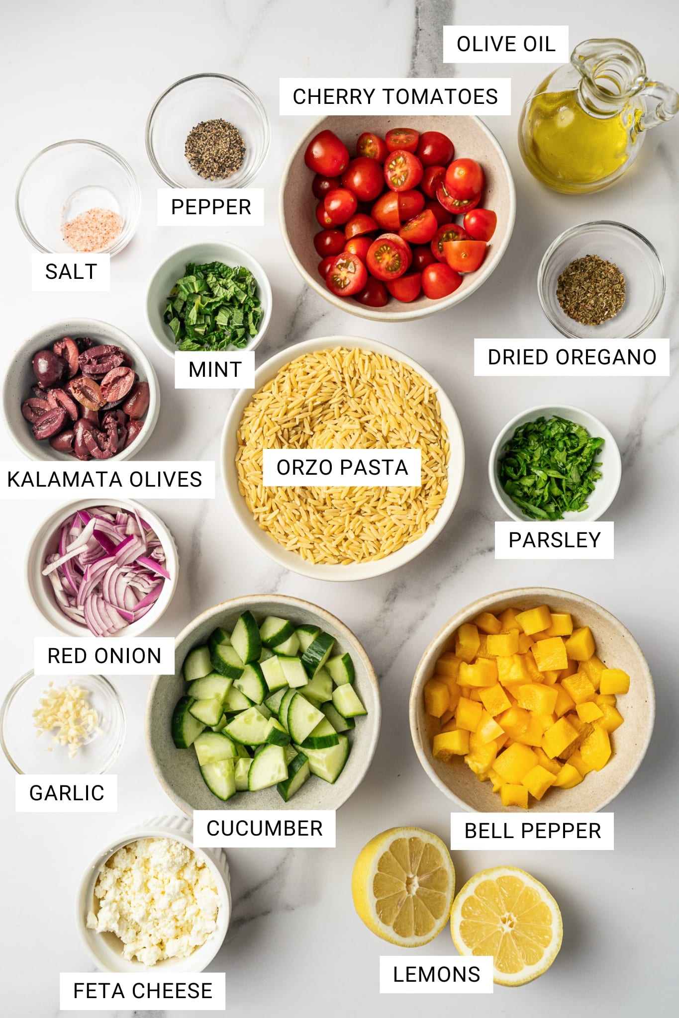 Overhead labeled ingredient image for Mediterranean orzo salad showing bowls of orzo pasta, cherry tomatoes, cucumber, yellow bell pepper, red onion, Kalamata olives, feta cheese, mint, parsley, garlic, dried oregano, salt, pepper, olive oil, and lemons on a white marble surface, with white text labels over each ingredient.