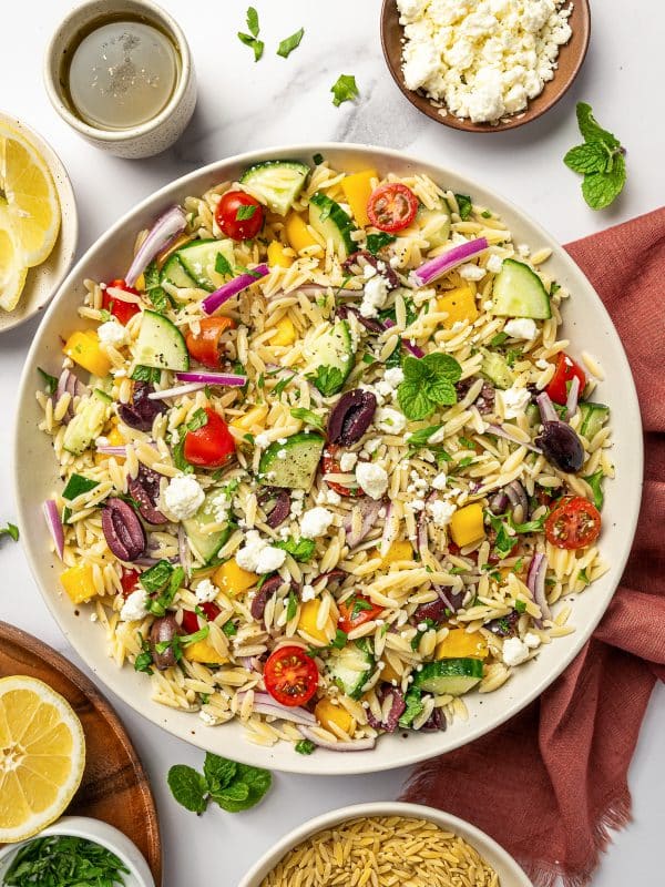 Overhead view of Mediterranean orzo salad in a large beige serving bowl with orzo, cucumber, cherry tomatoes, yellow bell pepper, red onion, Kalamata olives, crumbled feta, parsley, mint, and black pepper, surrounded by lemon halves, extra feta, olive oil, chopped parsley, dry orzo, and a rust-colored napkin on a white marble surface.