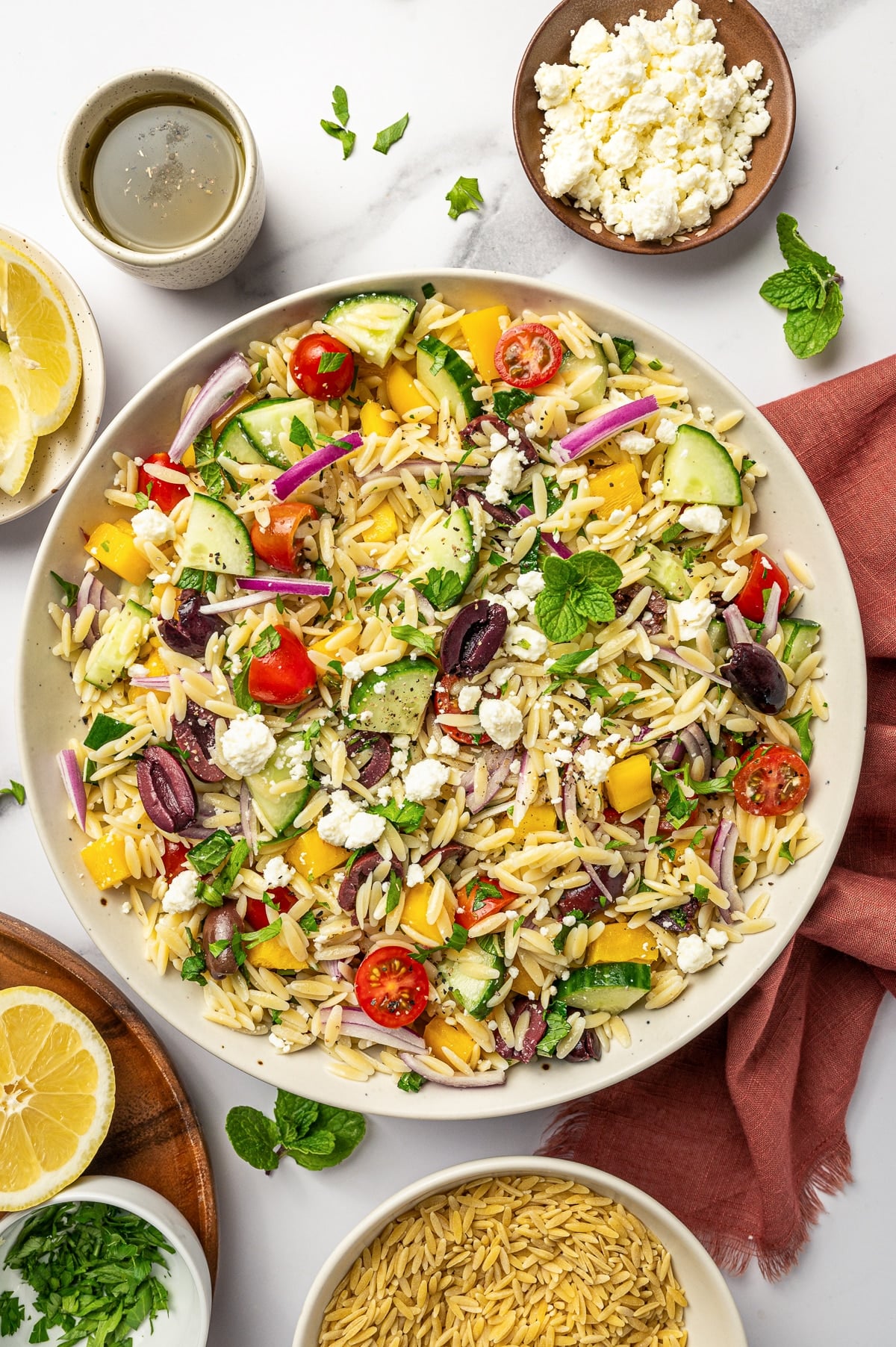 Overhead view of Mediterranean orzo salad in a large beige serving bowl with orzo, cucumber, cherry tomatoes, yellow bell pepper, red onion, Kalamata olives, crumbled feta, parsley, mint, and black pepper, surrounded by lemon halves, extra feta, olive oil, chopped parsley, dry orzo, and a rust-colored napkin on a white marble surface.