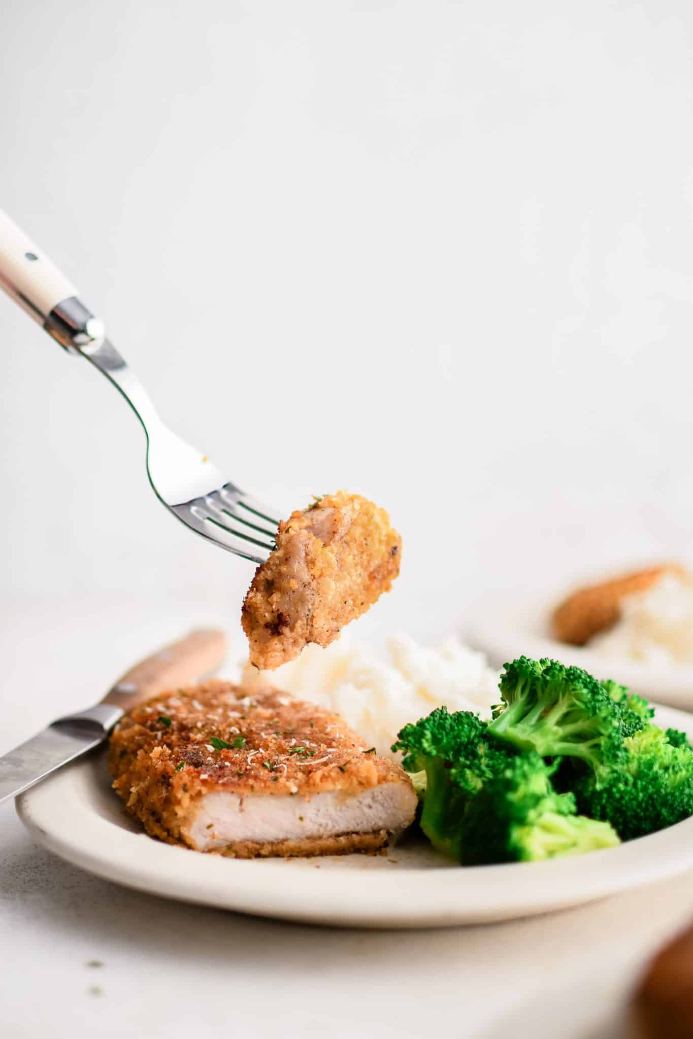Fork holding a bite of Parmesan crusted pork chop above a plate with sliced chop, mashed potatoes, and steamed broccoli.