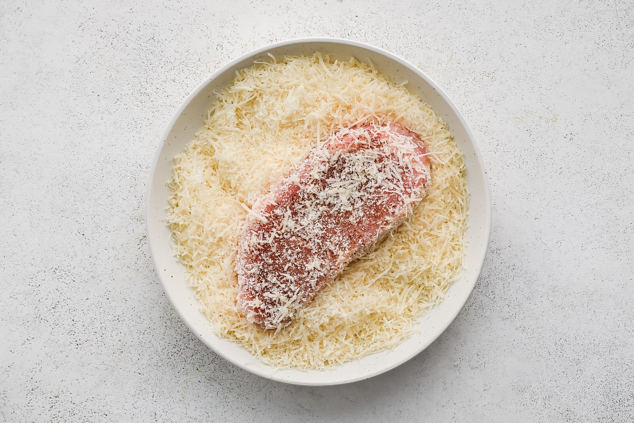 A single seasoned pork chop pressed into a shallow bowl filled with finely shredded Parmesan cheese.