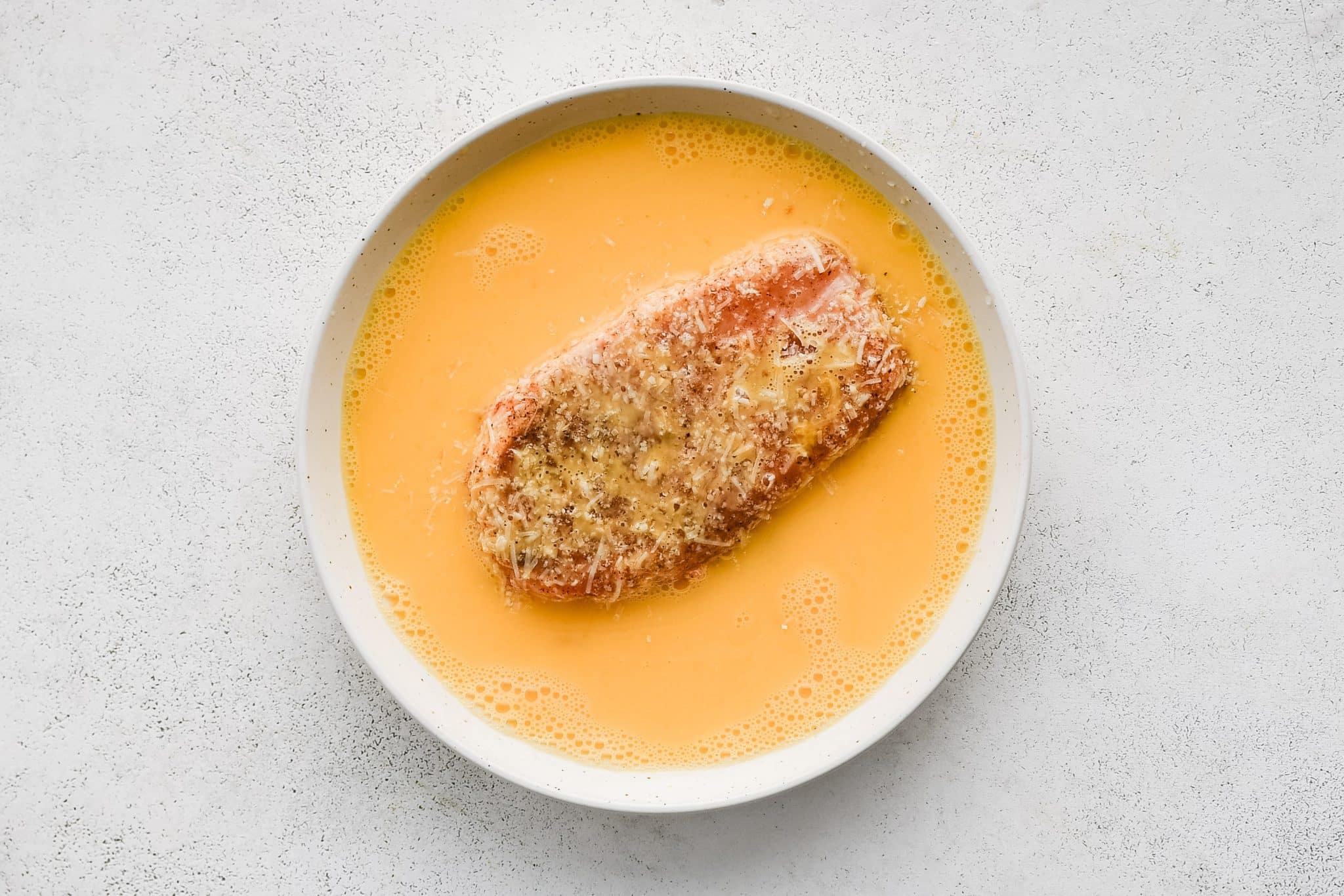 A Parmesan coated pork chop submerged in a bowl of beaten eggs to help the breadcrumb crust stick.
