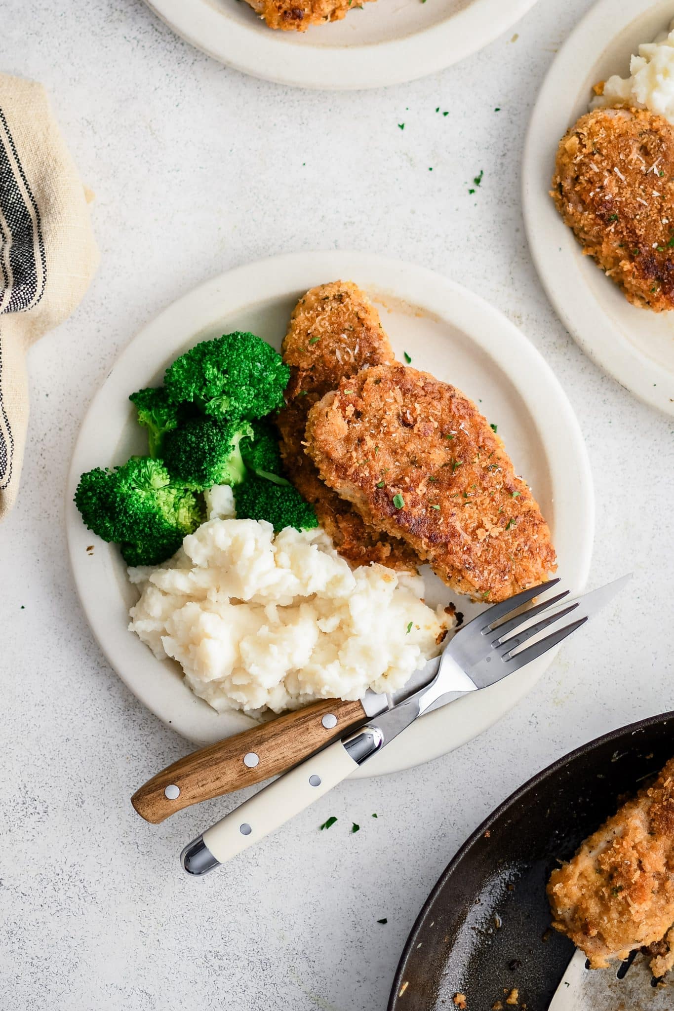 Overhead view of a plate with two Parmesan crusted pork chops, mashed potatoes, and broccoli with a fork and knife.