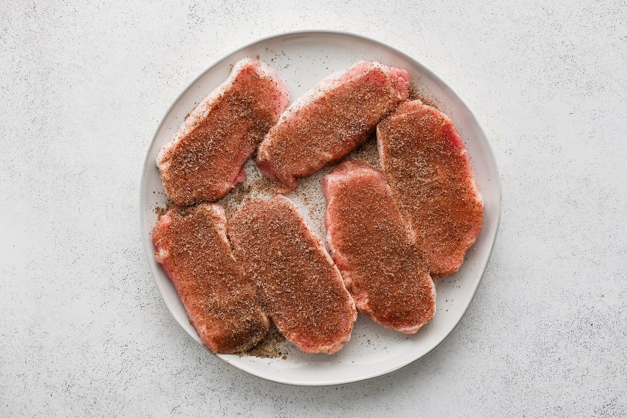 Six boneless pork chops on a white plate coated evenly with a blend of salt, pepper, paprika, garlic powder, and onion powder.