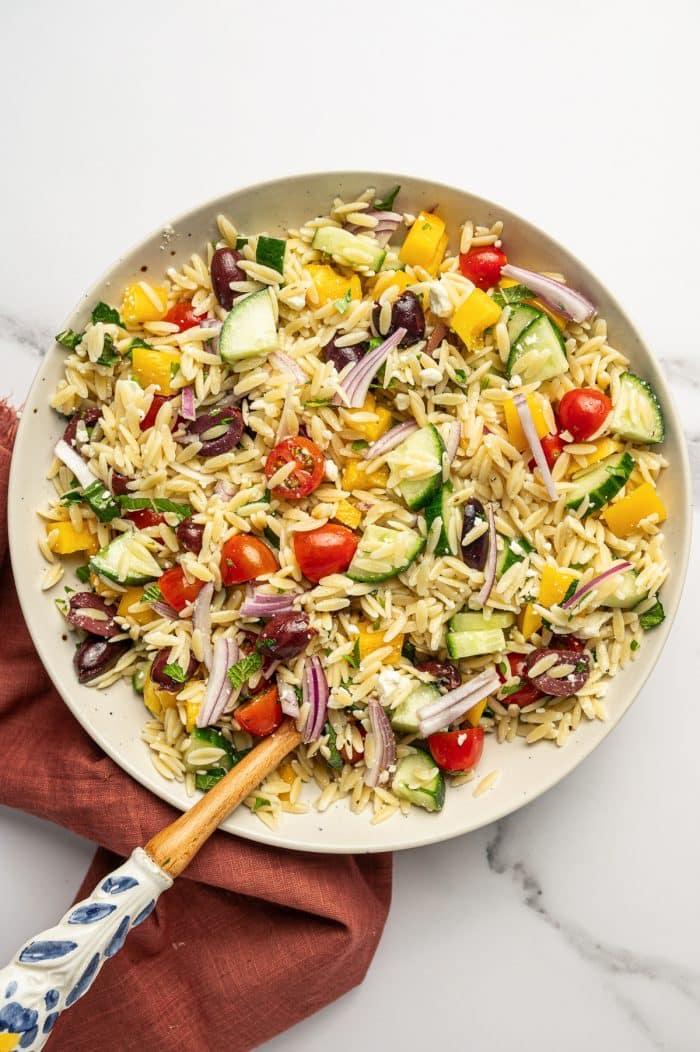 Tossed Mediterranean orzo salad in a large beige bowl with tender orzo pasta, cucumber, cherry tomatoes, yellow bell pepper, red onion, Kalamata olives, feta cheese, and fresh herbs, with a wooden spoon resting in the bowl and a rust-colored napkin underneath.