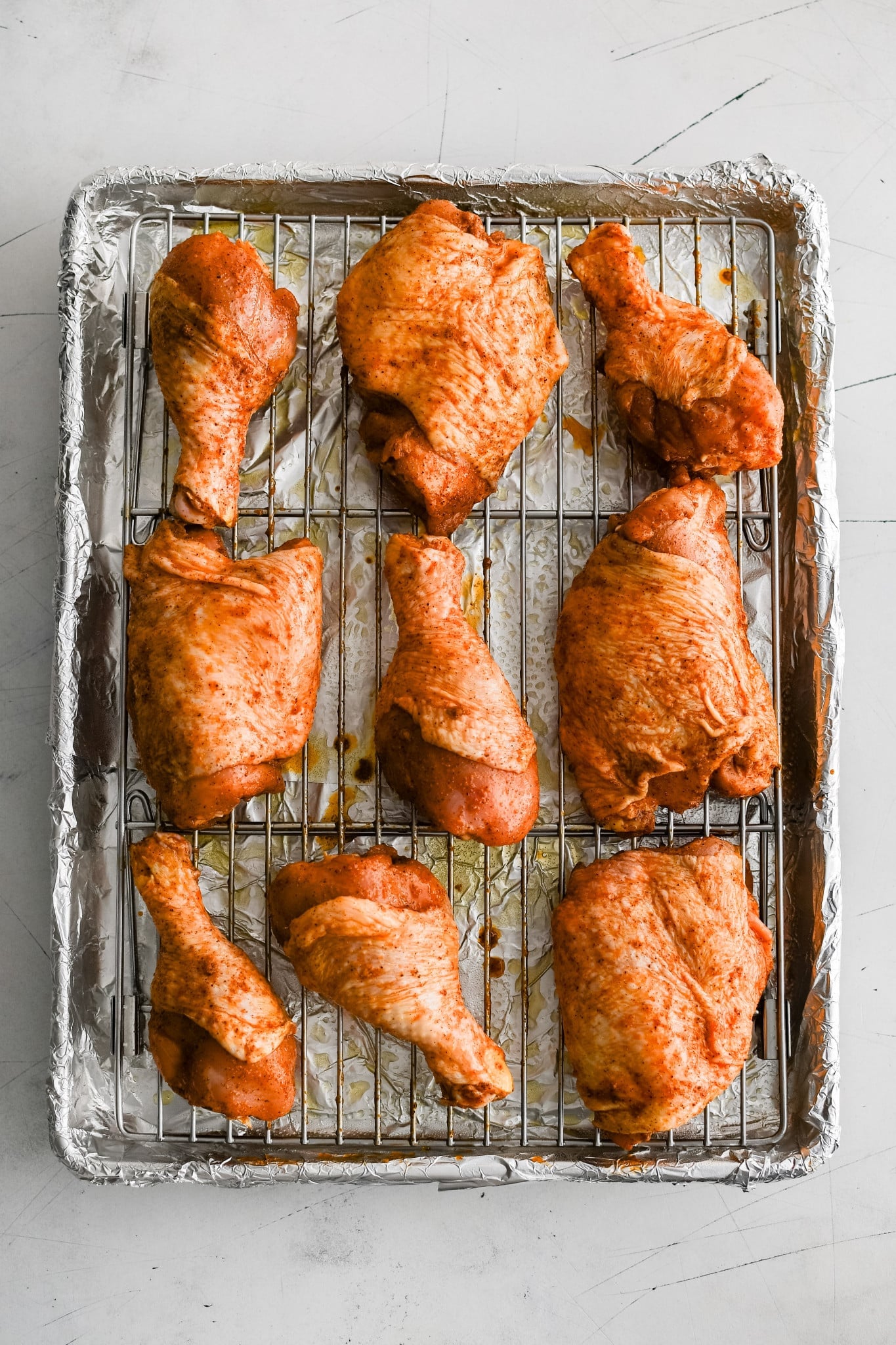 Combination of raw bone-in skin-on chicken thighs and drumsticks coated in dry rub on a cooling rack placed in a baking sheet.
