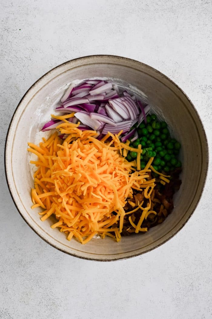 Shredded cheddar cheese, sliced red onion, green peas, and chopped bacon added to a large white mixing bowl filled with creamy ranch seasoning and sour cream dressing.