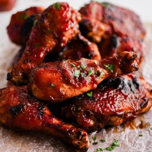 Cutting board lined with wax paper and piled with perfectly baked bone-in skin-on BBQ chicken thighs and drumsticks coated in sticky BBQ sauce and garnished with minced parsley.