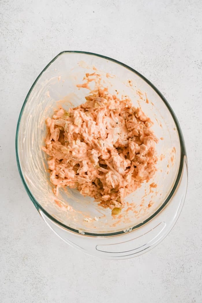 Large glass mixing bowl filled with shredded chicken mixed with creamy buffalo sauce.