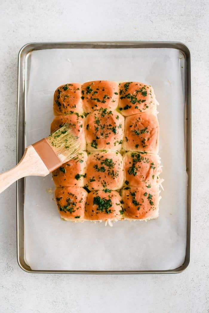 Brushing parsley infused melted butter over the tops of assembled buffalo chicken sliders made with sweet Hawaiian rolls, shredded chicken mixed in creamy buffalo sauce, and shredded mozzarella cheese.