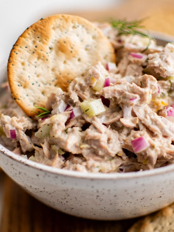 Single water cracker in a white bowl filled with creamy tuna salad presented on a cutting board surrounded by crackers and celery sticks.