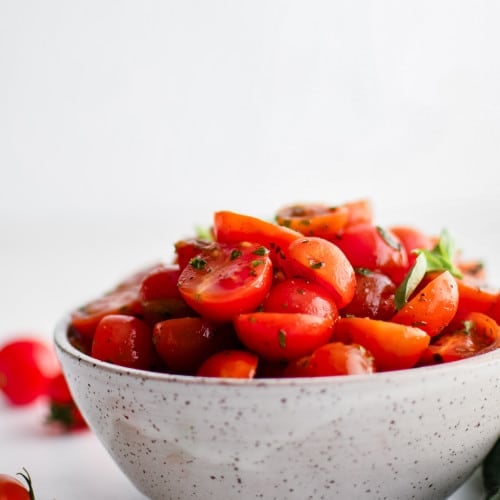 Cherry tomato salad in a rustic-style speckled bowl, styled with basil and whole cherry tomatoes around the base.