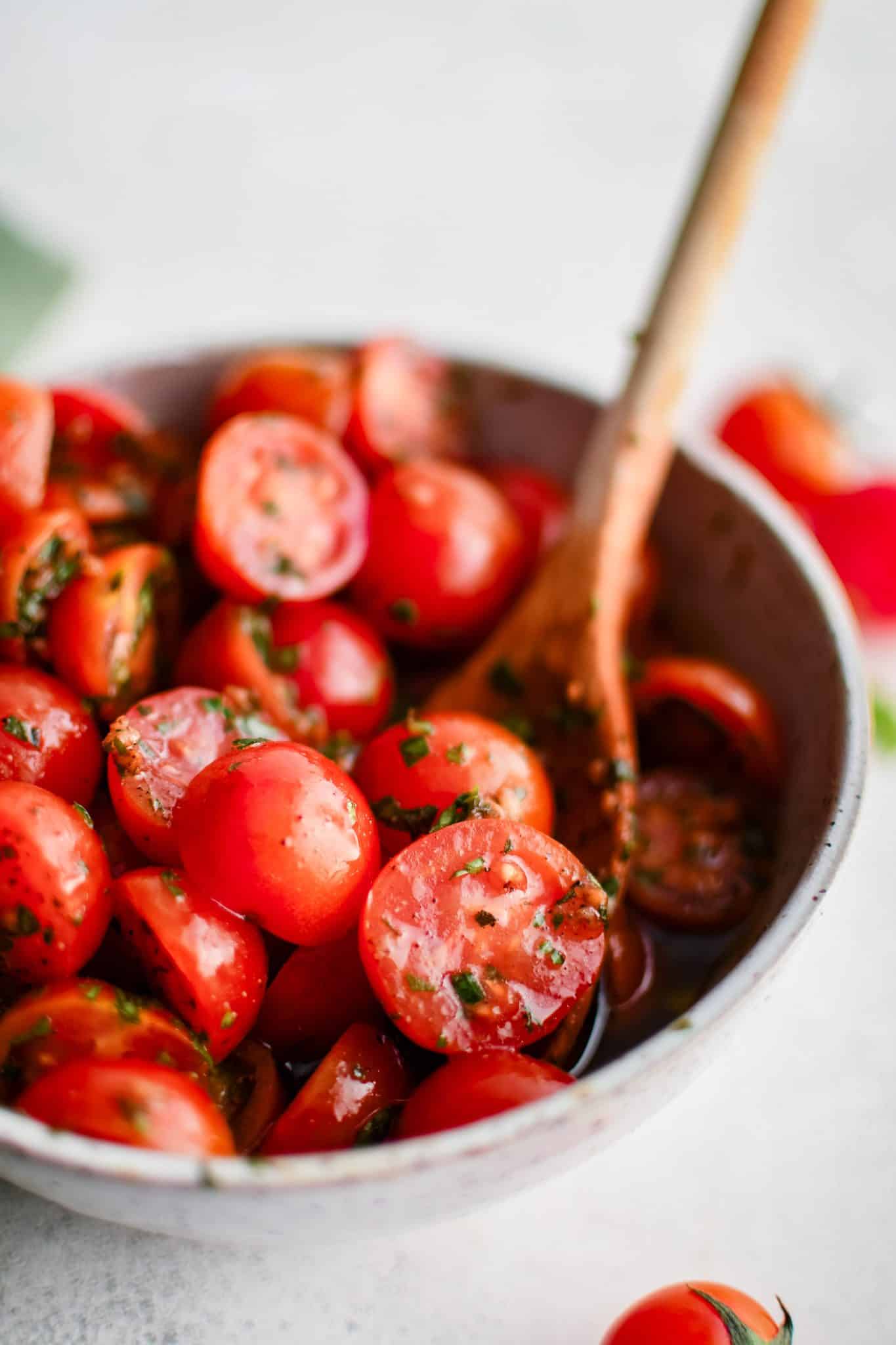 Large white salad bowl filled with halved cherry tomatoes tossed in olive oil, balsamic vinaigrette, garlic, salt, black pepper, and fresh herbs.