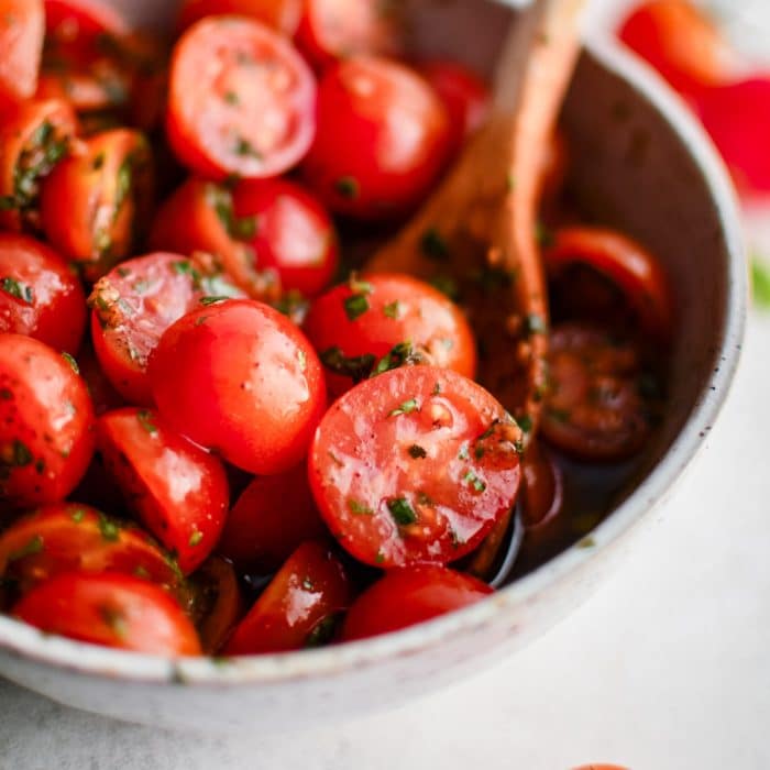 A full bowl of cherry tomato salad stirred with a wooden spoon, with glistening herbs and vibrant color.