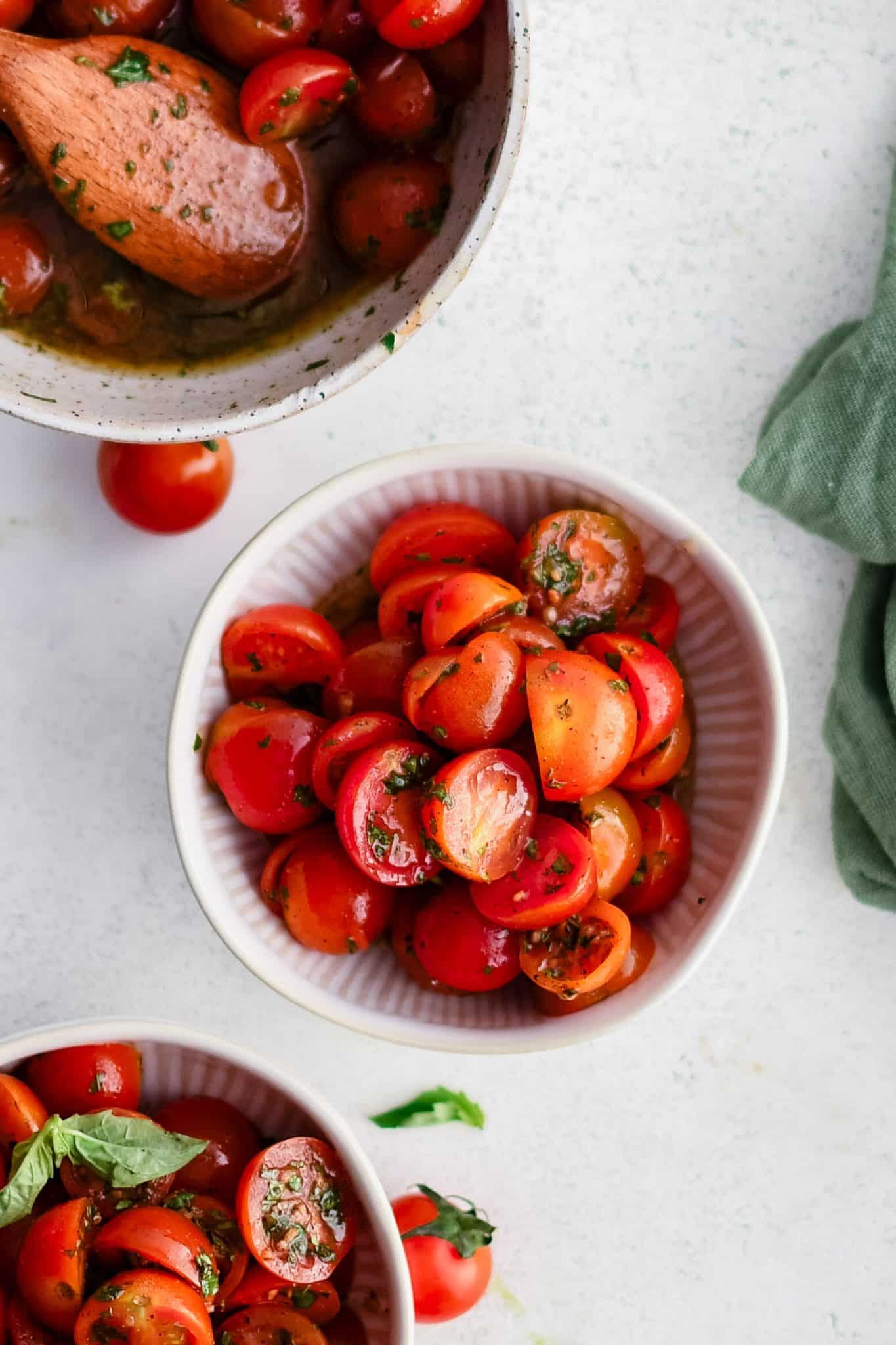 Small white serving bowls filled with halved cherry tomatoes tossed in olive oil, balsamic vinaigrette, garlic, salt, black pepper, and fresh herbs.