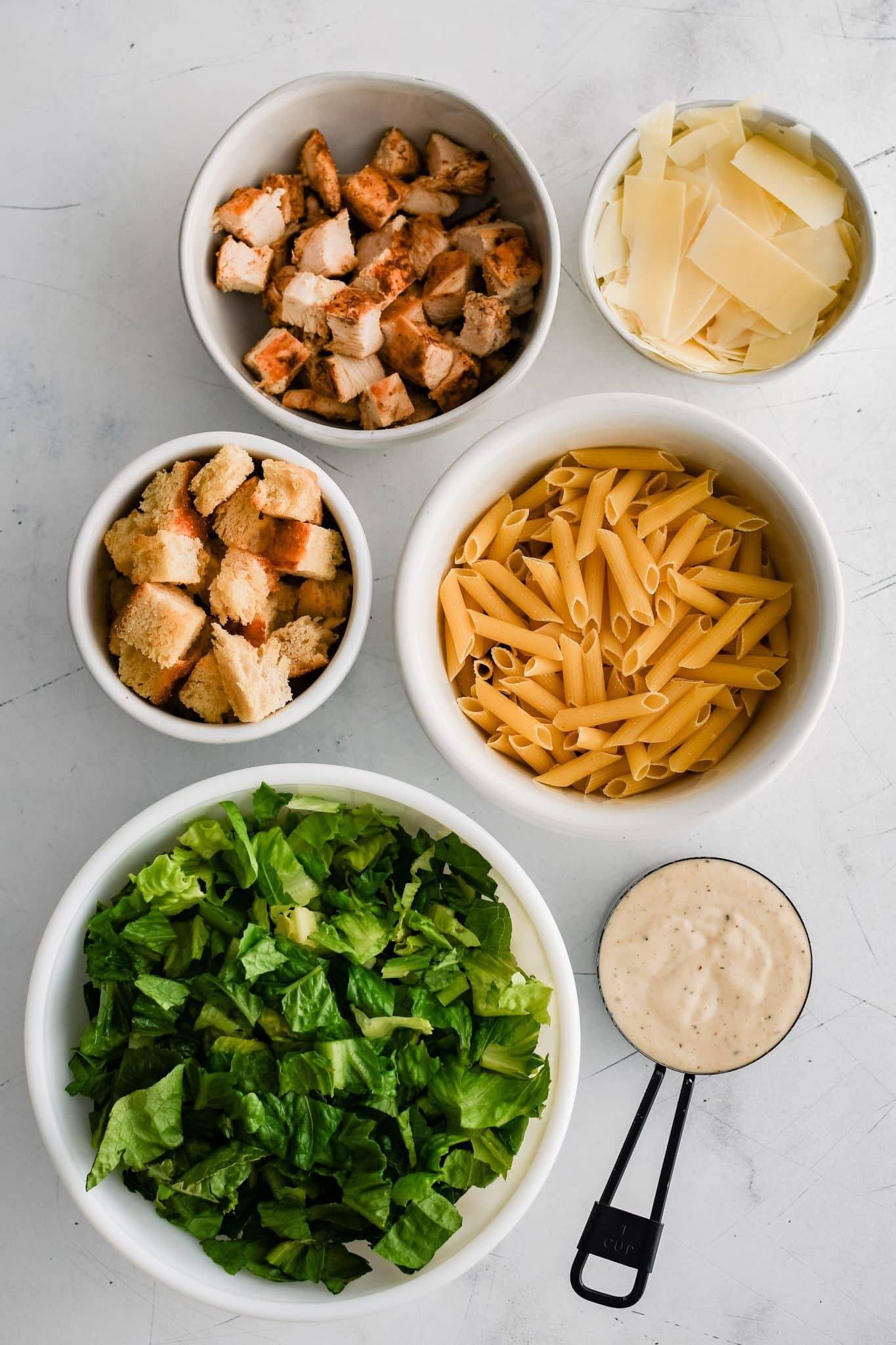 All of the ingredients for chicken Caesar pasta salad presented in individual measuring cups and ramekins.