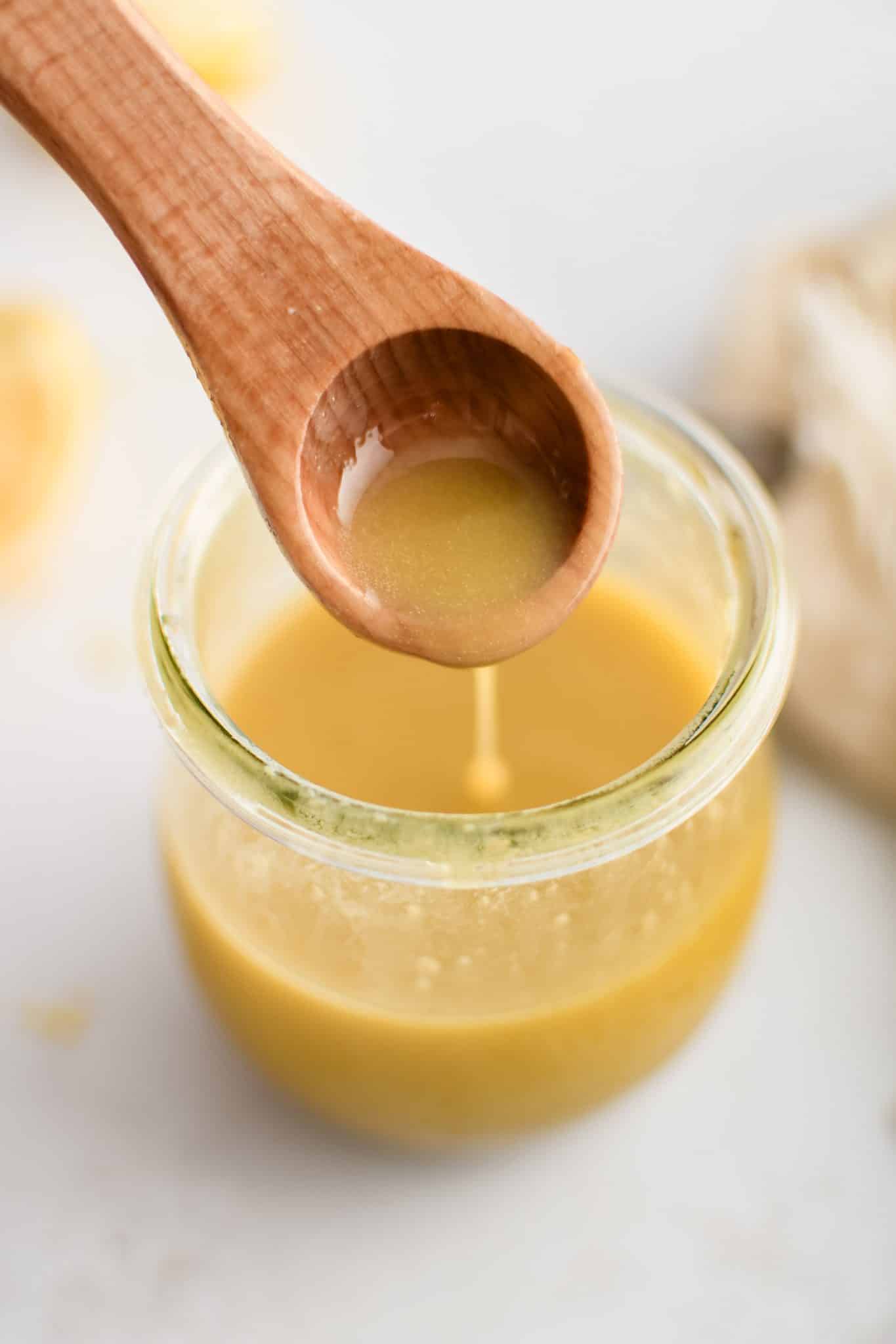 Small wood spoon filled with homemade Dijon vinaigrette hovering over a glass jar filled with vinaigrette.
