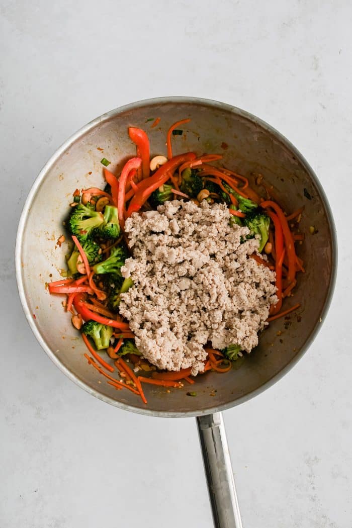 Pre-cooked ground chicken added to a large wok filled with cooking broccoli, bell pepper, carrots, and green onions.