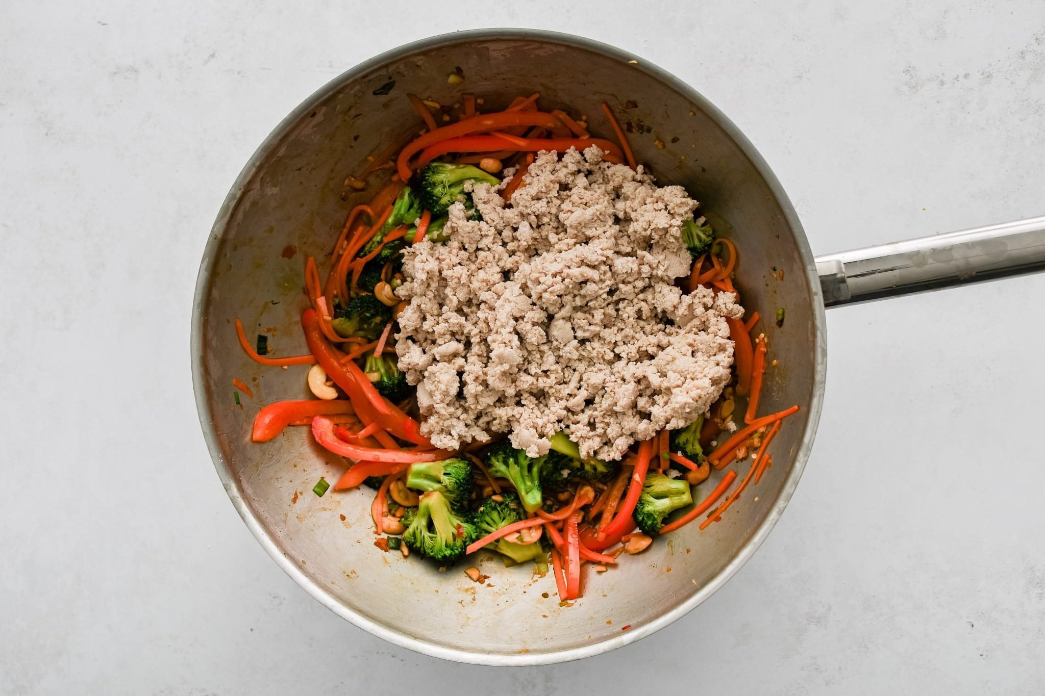 Pre-cooked ground chicken added to a large wok filled with cooking broccoli, bell pepper, carrots, and green onions.
