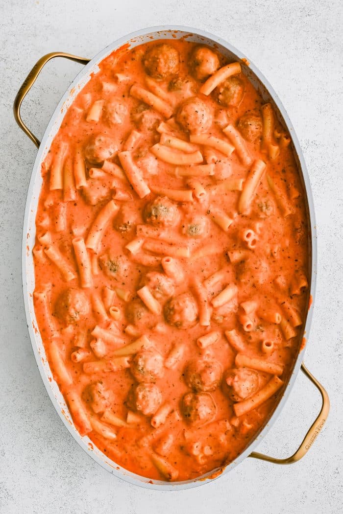 Par-baked meatball casserole with meatballs and ziti pasta in a creamy red sauce with mozzarella cheese cooking in a large oval-shaped casserole dish.