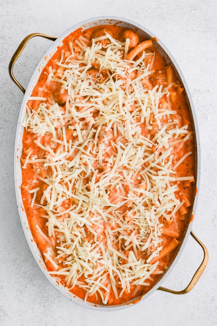 Mozzarella cheese sprinkled over par-baked meatball casserole with meatballs and ziti pasta in a creamy mozzarella red sauce cooking in a large oval-shaped casserole dish.