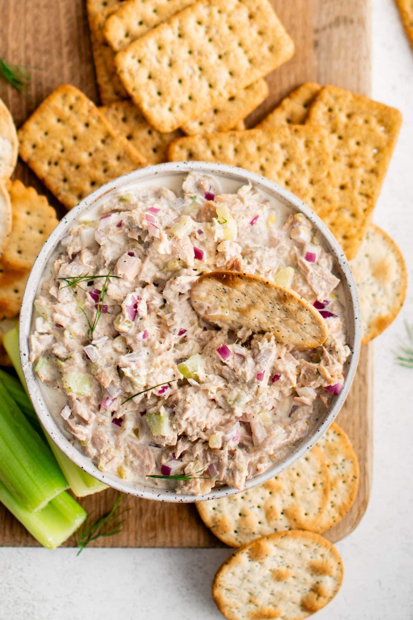 White bowl filled with homemade classic tuna salad on a large cutting board surrounded by whole grain crackers, celery sticks, water crackers, and butter crackers.
