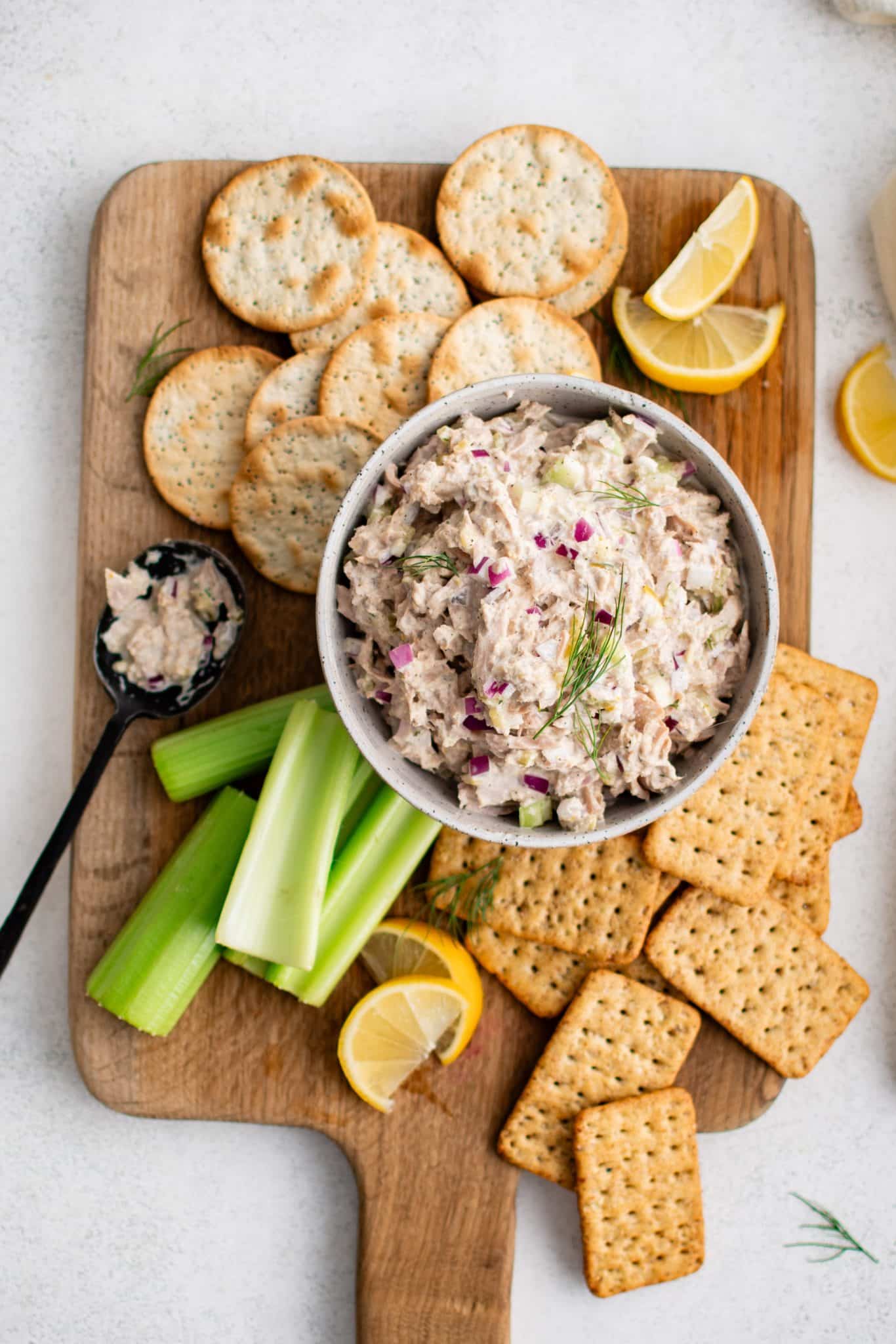 White bowl filled with homemade classic tuna salad on a large cutting board surrounded by whole grain crackers, celery sticks, water crackers, and butter crackers.