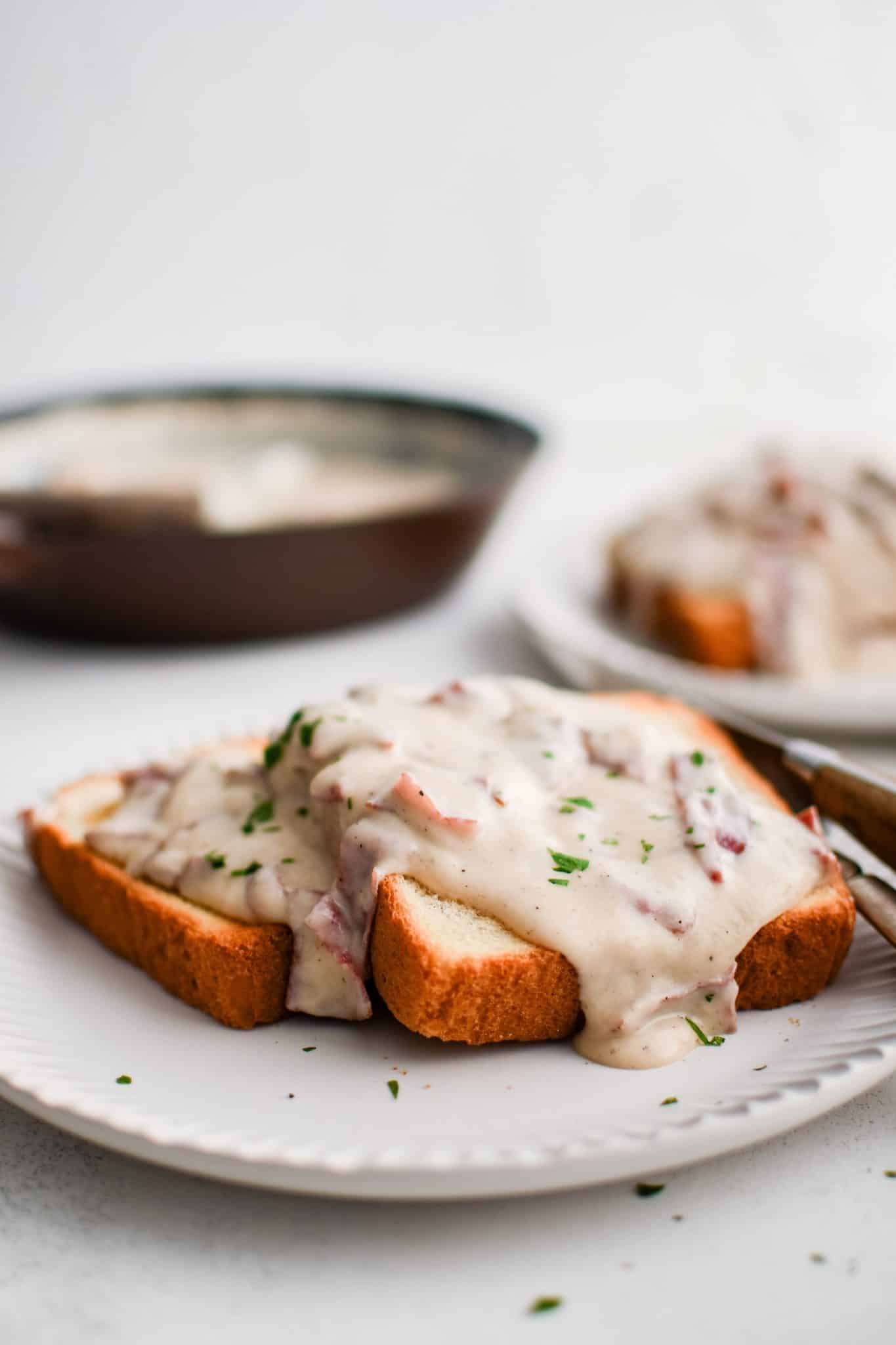 Two slices of golden toasted bread on a white plate topped with a generous serving of creamed chipped beef spilled over the sides and garnished with fresh parsley with the cast iron skillet filled with the remain creamed chipped beef in the background.