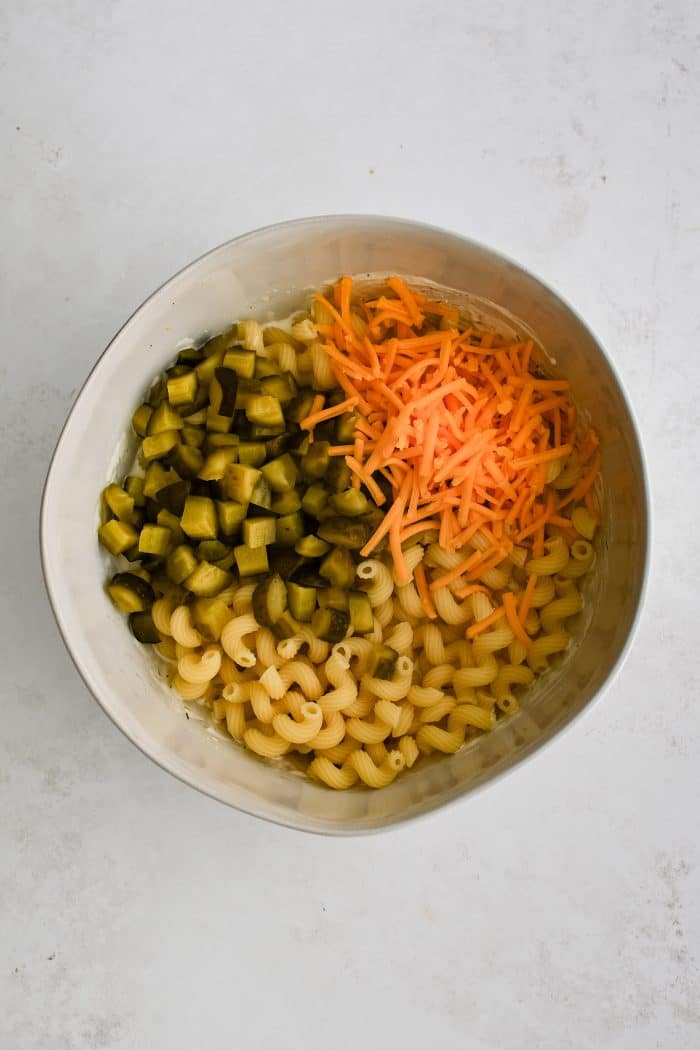 Large white mixing bowl filled with cooked Cavatappi pasta, chopped dill pickles, and shredded cheddar cheese.