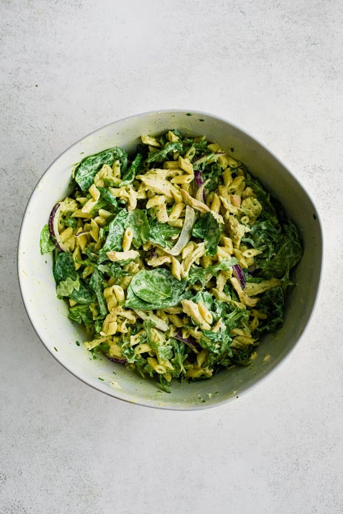Large white mixing bowl filled with creamy green goddess pasta salad made with cooked pasta, shredded chicken, sliced red onion, spinach, arugula, and give mixed with green goddess dressing.