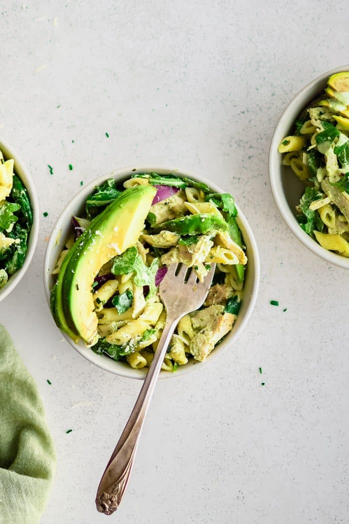 Three small serving bowls (two of the bowls only partially in image) filled with tossed together green goddess pasta salad and topped with a large slice of fresh avocado.