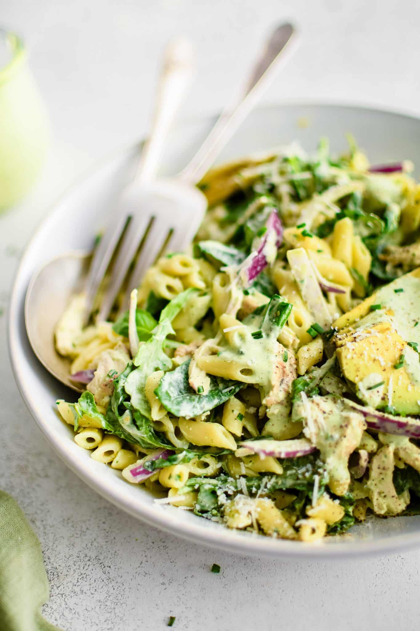 Large white serving bowl filled with creamy green goddess pasta salad made with short penne pasta, shredded chicken, sliced red onion, fresh spinach, arugula, and avocado all tossed in homemade green goddess dressing.